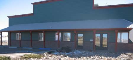 A single-story commercial building with green siding, red trim, several windows, and a covered entrance facing a rocky landscaped area.