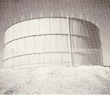 Large cylindrical water tank on a dirt mound, surrounded by a chain-link fence with barbed wire at the top, photographed in black and white.