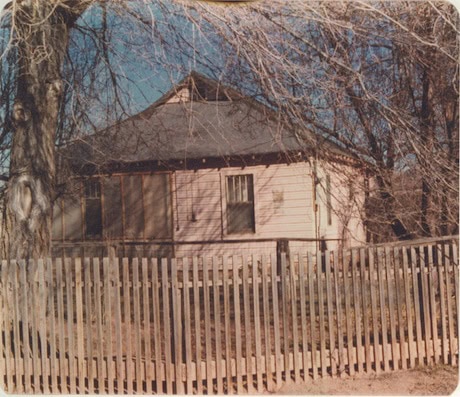 A small, old wooden house with a fenced yard, surrounded by leafless trees under a clear sky.
