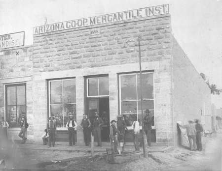 Black-and-white photo of people standing outside a stone building labeled "Arizona Coop Mercantile Inst." The scene appears historic, with individuals in period clothing.