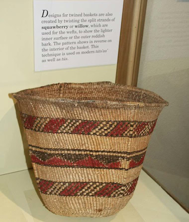 A woven basket with red and black geometric patterns sits on display, accompanied by an informational sign about its construction materials and techniques.