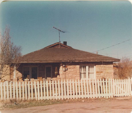 A single-story stone house with a dark roof, white picket fence, and an old television antenna on top, photographed on a clear day.
