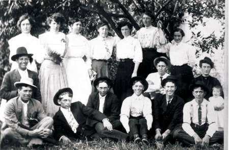 A black and white photo of fifteen adults and one child, posed outdoors in two rows under a tree. Most are wearing early 20th-century clothing and hats.