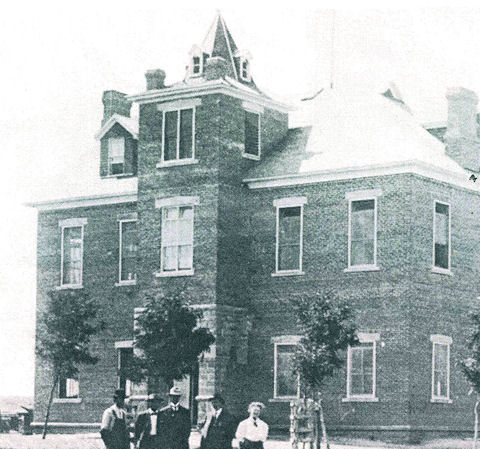 A group of people stands in front of a large, two-story brick building with a tower and multiple chimneys.