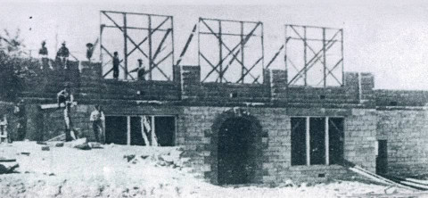 Black and white photo of a partially constructed stone building with workers standing on scaffolding above the first story.
