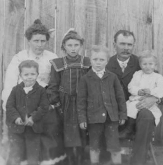 Black-and-white photo of a family with two adults and four children posing in front of a wooden wall, dressed in early 20th-century clothing.