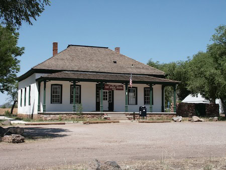 A historic one-story building with a hipped roof, white walls, green trim, and a small sign above the entrance, situated in a rural area with trees and gravel surroundings.