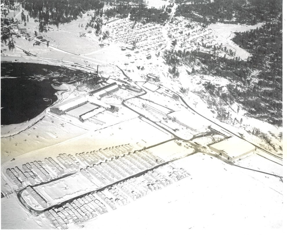 Aerial view of a snow-covered industrial facility with several large buildings, roads, and stacks of materials near a body of water, surrounded by forested areas.