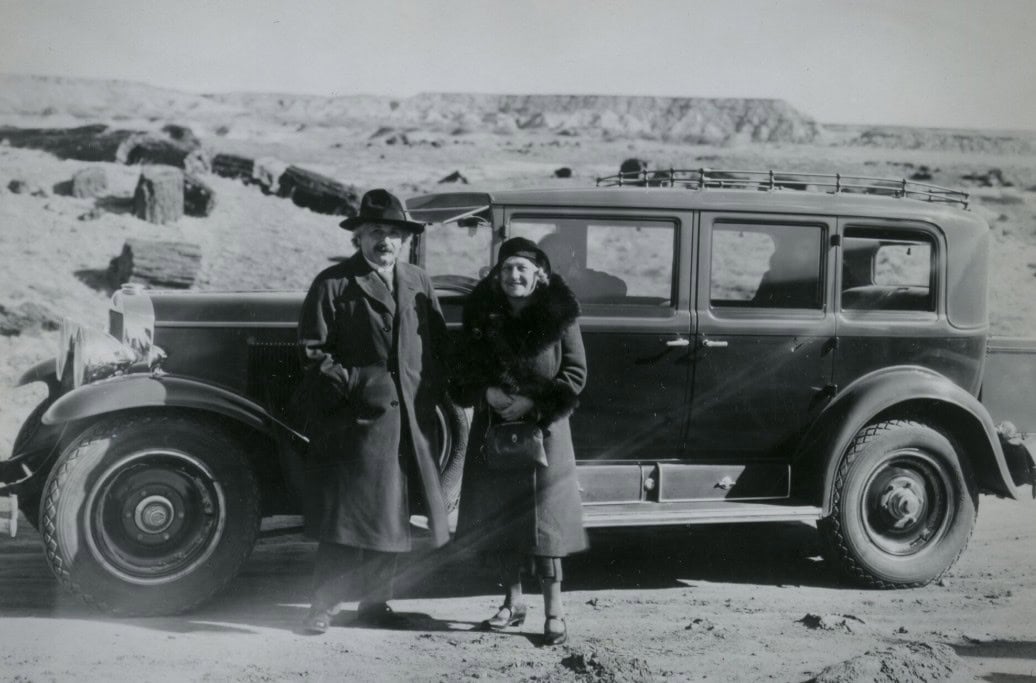 A man and a woman in coats stand in front of a vintage car in a barren, rocky landscape.