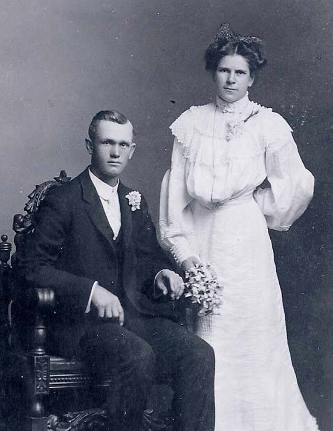A black-and-white vintage portrait of a man in a suit seated on a chair next to a woman in a white dress holding flowers, likely from the early 20th century.