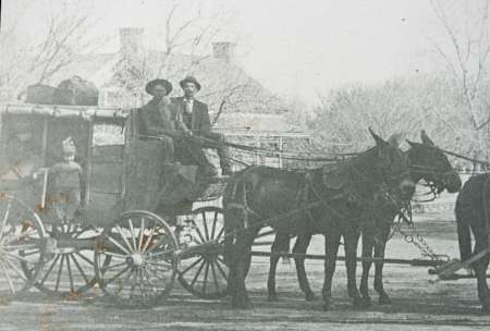 Black-and-white photo of a horse-drawn carriage with two men seated at the front, a child standing beside, and trees and a house in the background.