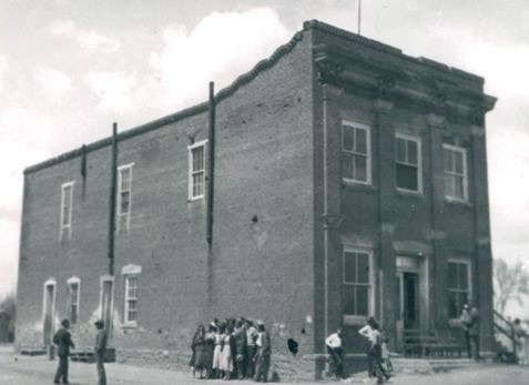 Black and white photo of a two-story brick building with groups of people gathered near the entrance and along the side.