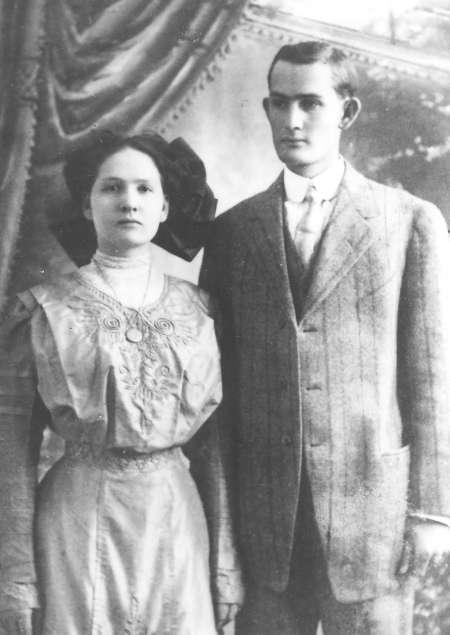 A black and white portrait of a young woman and man standing side by side, dressed in early 20th century formal attire, in front of a studio backdrop.