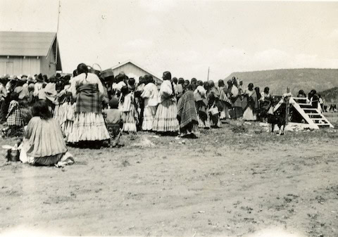 A group of people in traditional clothing gather outdoors near wooden buildings, with hills visible in the background on a sunny day.