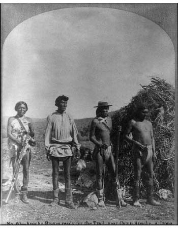 Four Apache men stand in front of a brush shelter holding rifles, dressed in traditional and Western-style clothing; arid landscape in the background.