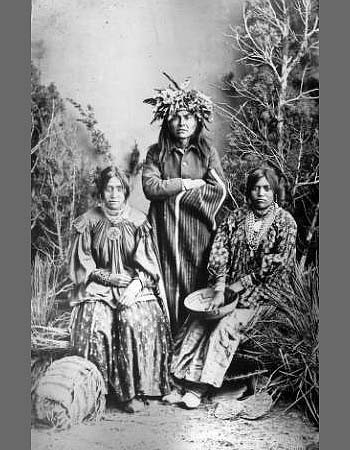 Three Indigenous people pose for a black-and-white portrait outdoors; one stands wearing a headpiece and robe, while two sit in patterned clothing, one holding a shallow basket.