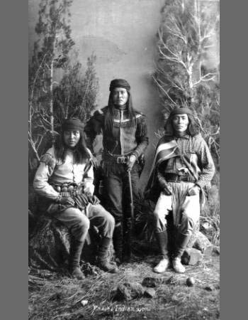 Three Apache men wearing traditional clothing pose for a studio portrait in front of a painted forest backdrop, with trees and rocks at their feet.