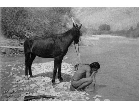A person kneels by a river washing their face while a horse stands nearby on the rocky shore.