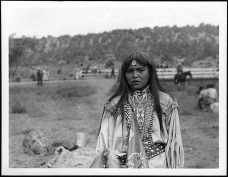 A Native American woman stands outdoors wearing traditional clothing and jewelry, with a rural landscape and several people in the background.