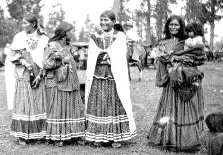 Four Native American women in traditional dress stand outdoors; one woman holds a child. Trees and people on horseback are visible in the background.