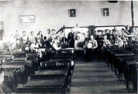 A black-and-white photo of a classroom with rows of empty desks in the foreground and a group of people, some with musical instruments, gathered at the back of the room.