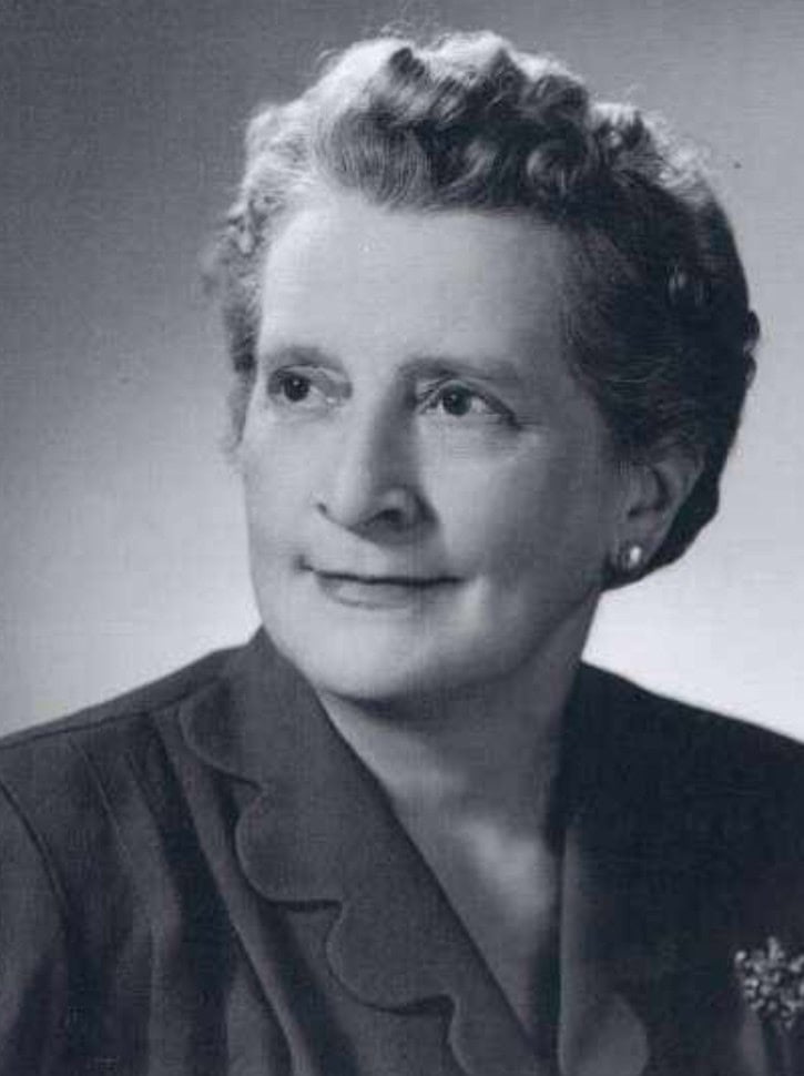 Black and white portrait of an older woman with short, wavy hair, wearing a suit jacket with scalloped edges and a brooch, looking slightly to the side.