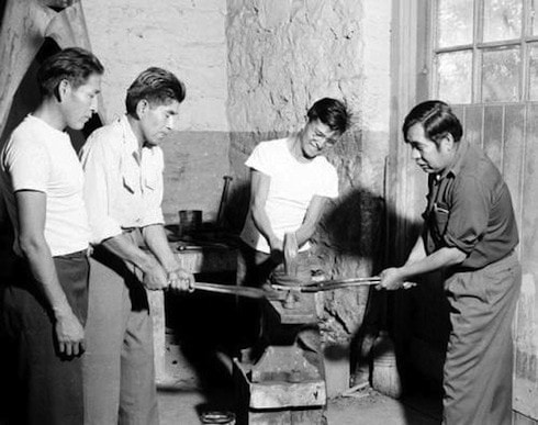 Four men work together in a workshop, using tools to shape metal on an anvil; two observe while two operate the equipment.
