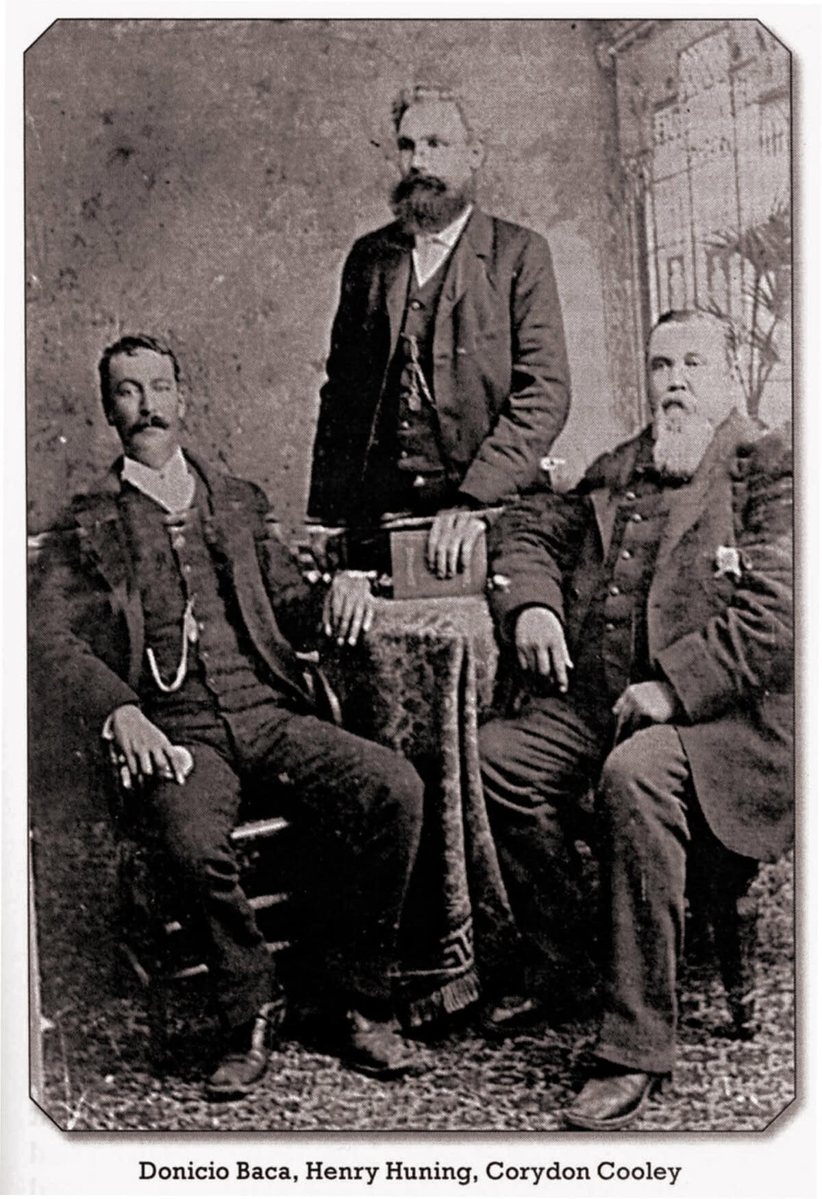 Three men in formal 19th-century attire pose for a portrait; two are seated and one stands behind a small table.