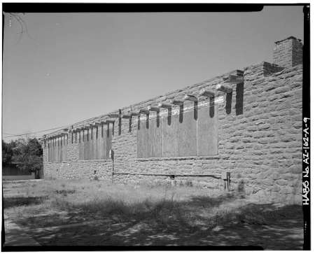 Black-and-white photo of a long, single-story stone building with boarded-up windows and exposed beams under the roofline, surrounded by dry grass.
