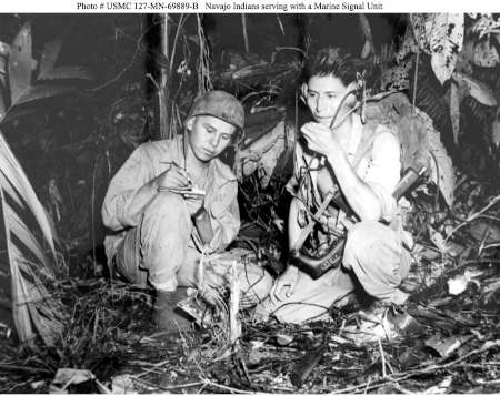 Two soldiers in military uniforms sit in dense foliage; one writes in a notebook while the other uses a radio headset and holds a microphone.