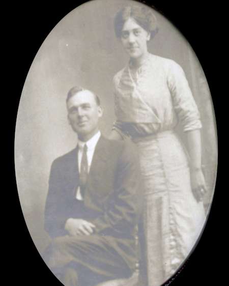 A sepia-toned oval portrait of a man in a suit seated next to a woman standing in a long dress, both facing the camera.