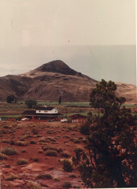 A white house with a red shed sits in a dry, shrub-covered landscape, with a large rocky hill or small mountain in the background under a cloudy sky.