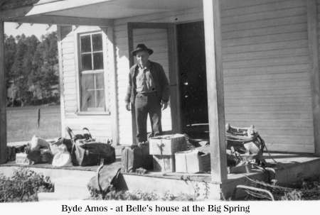 A man stands on the porch of a house with many bags and boxes scattered in front; trees and grass are visible in the background.
