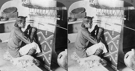 A person sits on a mat, weaving a patterned textile on a vertical loom, with pottery vessels nearby.