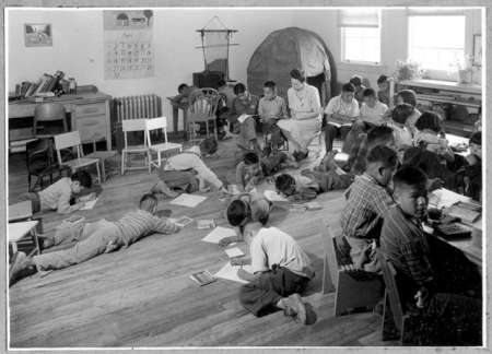 Black-and-white photo of children in a classroom, some seated at desks and others lying or sitting on the floor, writing or drawing, with a teacher present in the background.