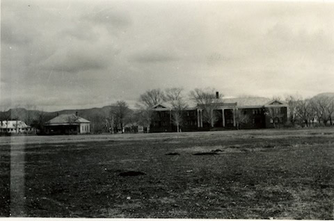 Black and white photo of large, columned building and two smaller structures on a barren field with trees and mountains in the background.