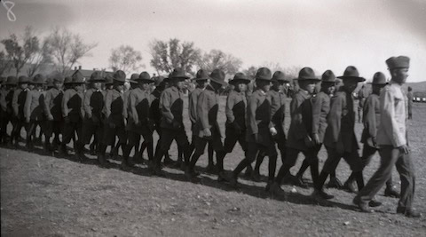A group of soldiers in uniform march in formation outdoors, led by an officer; trees and open land are visible in the background.