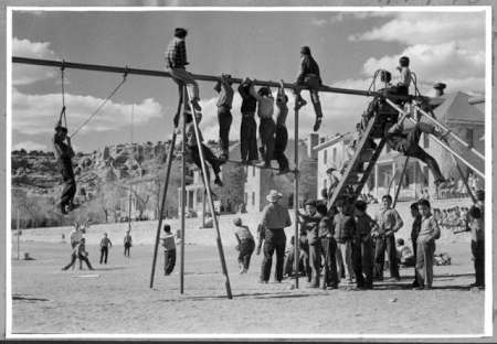 Children and adults gather around a playground with swings, a slide, and a metal climbing bar on a sunny day; buildings and rocky hills are visible in the background.