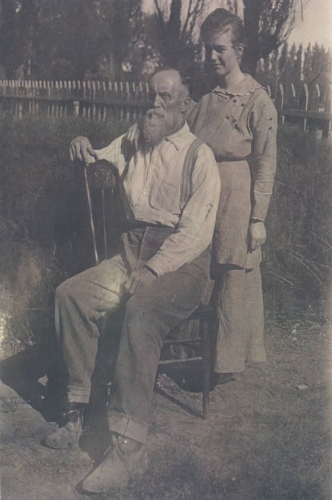 An elderly man with a long beard sits on a chair outdoors while a woman stands beside him, both dressed in early 20th-century clothing, with a wooden fence and trees in the background.