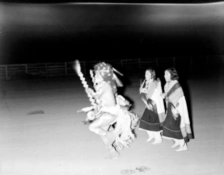 A person in traditional attire dances with a decorated staff while three women in shawls stand behind, outdoors at night.