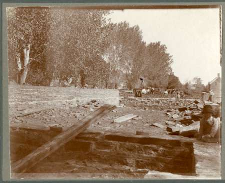 Sepia-toned photo of people working amid scattered debris and unfinished stone walls, with trees and a house visible in the background.