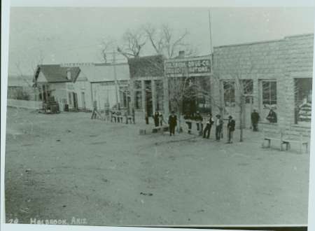 Black and white photo of a dirt street lined with buildings and storefronts; several people are standing or sitting along the sidewalk, with leafless trees in front.