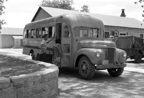 A vintage school bus is parked outside a building as two children board, carrying bags, with another vehicle visible in the background.