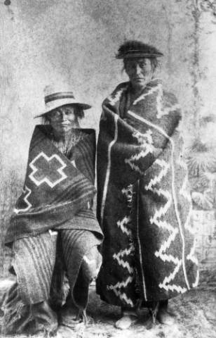Two Native American individuals in patterned blankets pose for a black-and-white photograph; one is seated and the other is standing, both wearing hats.