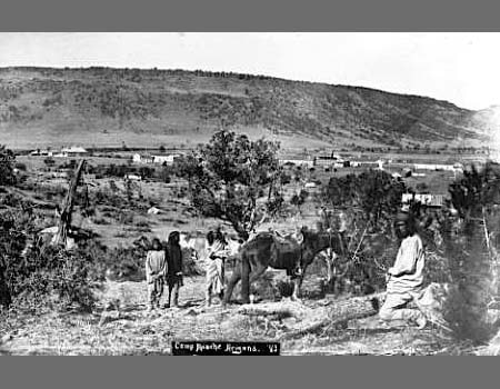 Black and white photo of several people and a horse in a rural landscape with scattered trees and buildings in the background near a hillside.
