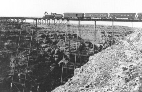 A train crosses a tall, narrow bridge spanning a rocky canyon, with steep cliffs visible on both sides.