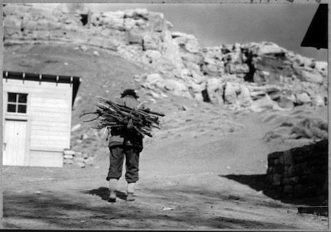 A person carrying a bundle of firewood on their back walks along a dirt path near a building, with rocky hills in the background.