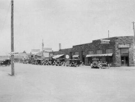 Black and white photo of a small town street with vintage cars parked in front of stone buildings, including a theater and pharmacy.