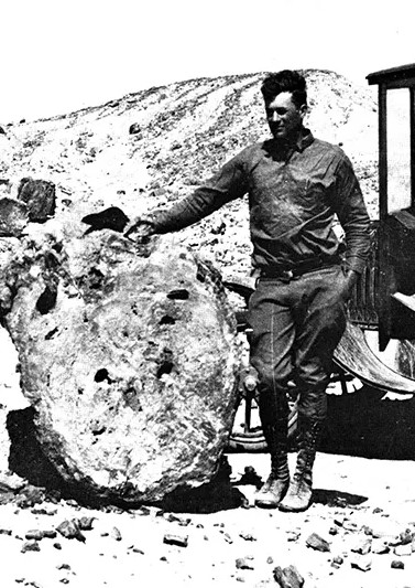 A man stands outdoors beside a large, rough meteorite resting on the ground, with rocky terrain and part of a vehicle visible in the background.