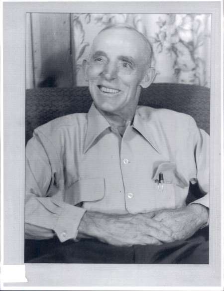 Elderly man with a bald head and a large smile sits in an armchair, wearing a light-colored button-up shirt with pens in the pocket.